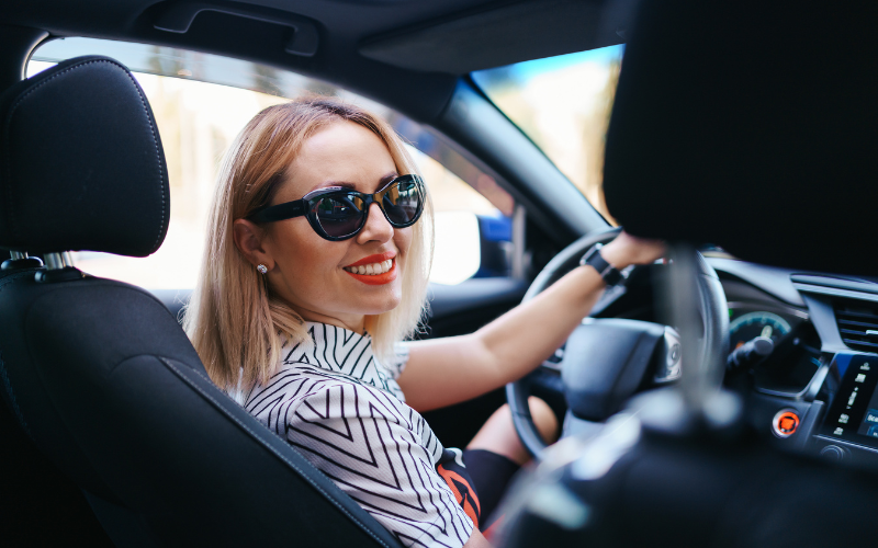 Una mujer rubia con gafas de sol oscuras sonríe al conductor mientras está en el coche. Ella lleva una camisa de rayas horizontales y tiene el cabello corto y rubio. Sus ojos están fijos en la persona que conduce, y su sonrisa es cálida y amigable. Las gafas de sol oscuras añaden un toque de misterio y sofisticación. El coche es de color azul y el fondo es una calle con árboles y edificios. Esta imagen captura un momento feliz y relajado en la vida de esta mujer.